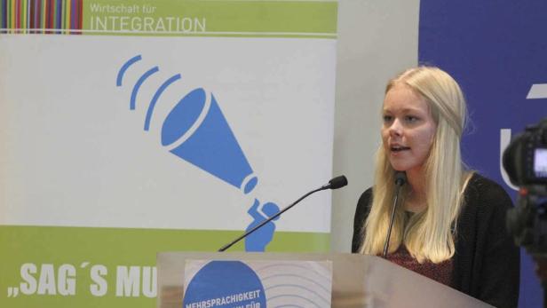 Eine blonde Frau spricht an einem Rednerpult vor einem Banner der Initiative „Wirtschaft für Integration“.