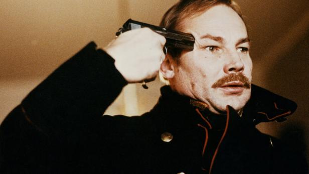 Ein Mann im Militärmantel hält sich eine Pistole an die Schläfe.