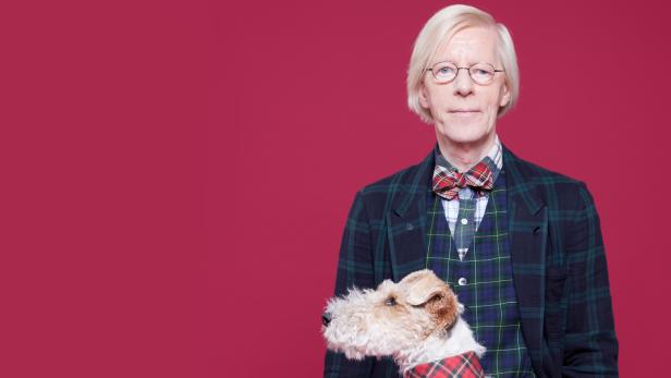 Ein Mann mit blonden Haaren und Brille posiert mit seinem Terrier vor einem roten Hintergrund.