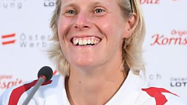 Eine lächelnde Sportlerin im österreichischen Nationaltrikot vor einem Werbehintergrund.