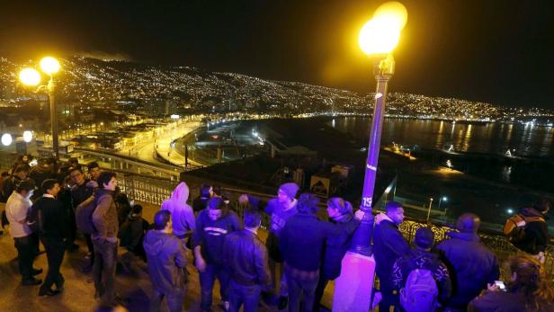 Eine Menschengruppe blickt nachts auf eine beleuchtete Stadt am Meer.