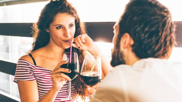 Eine Frau mit lockigem Haar und ein bärtiger Mann sitzen sich gegenüber und trinken ein Getränk aus Gläsern mit Strohhalmen.