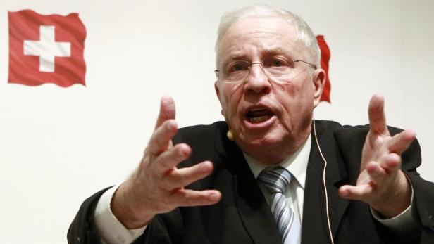 Ein Mann mit Brille gestikuliert vor einer Schweizer Flagge.