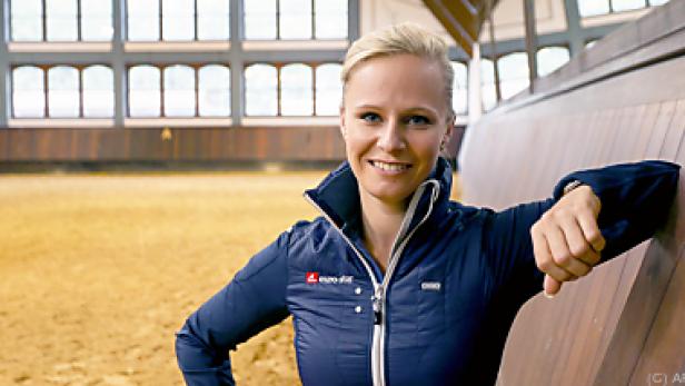 Eine blonde Frau lächelt, während sie in einer Reithalle an einer Holzwand lehnt.