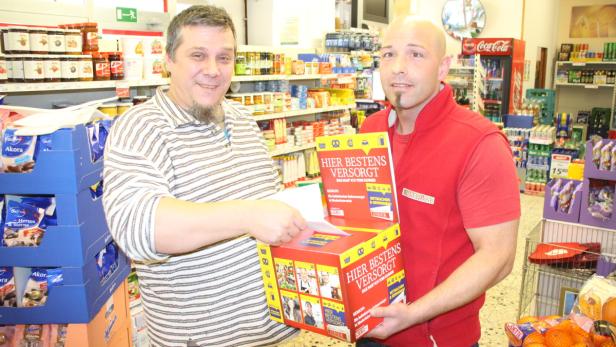 Zwei Männer stehen in einem Supermarkt neben einem Loskasten mit der Aufschrift „Hier bestens versorgt“.