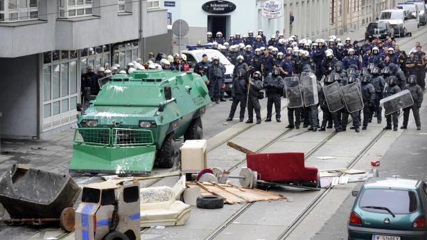 Ein grüner Panzerwagen steht vor einer Reihe Polizisten mit Schilden auf einer Straße mit Barrikaden.