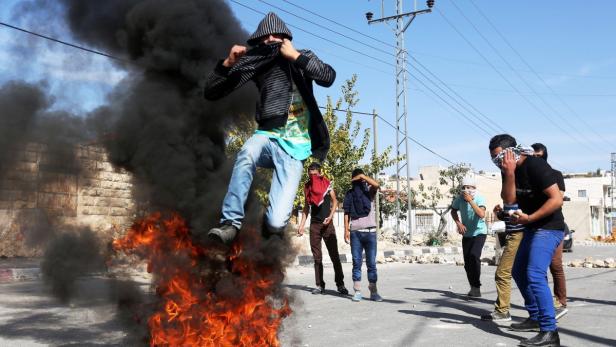 Ein Mann springt über ein Feuer, während andere mit Tüchern ihre Gesichter bedecken.