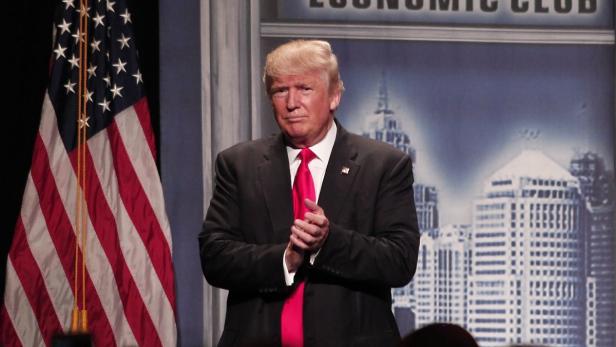 Donald Trump steht vor einer US-Flagge und einem Hintergrund mit einer Skyline.