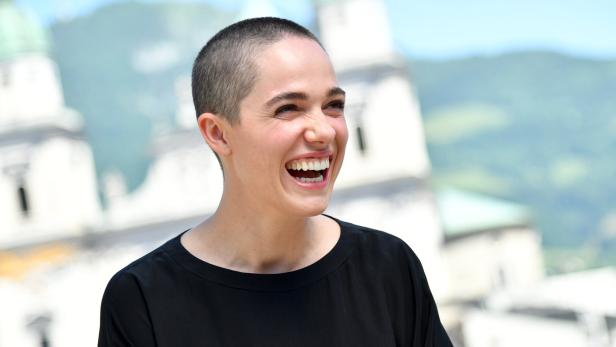 Eine lachende Frau mit kurz geschorenen Haaren vor einem Gebäude.