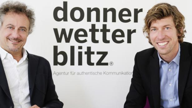 Zwei Männer in Anzügen posieren vor dem Logo der Agentur „donnerwetterblitz®“.