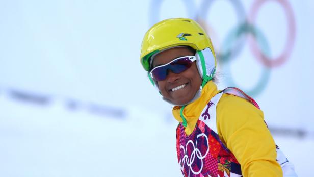 Eine lächelnde Skifahrerin mit gelbem Helm und Brille vor dem Hintergrund der Olympischen Ringe.