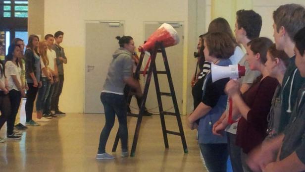 Eine Gruppe von Menschen steht in einem Raum, während eine Frau eine Leiter mit einem großen Megaphon schüttelt.