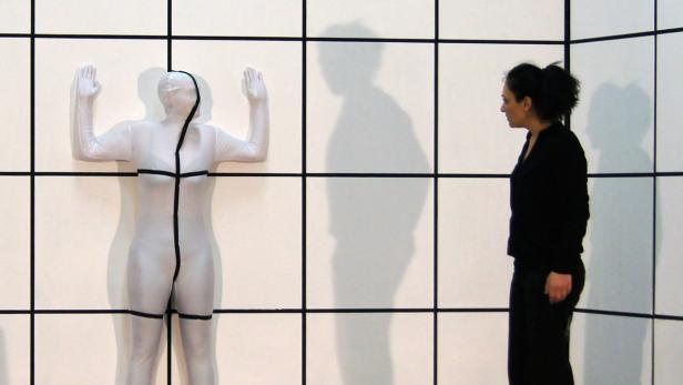 Eine Frau betrachtet eine an eine Wand gelehnte, in einen Ganzkörperanzug gekleidete Person.