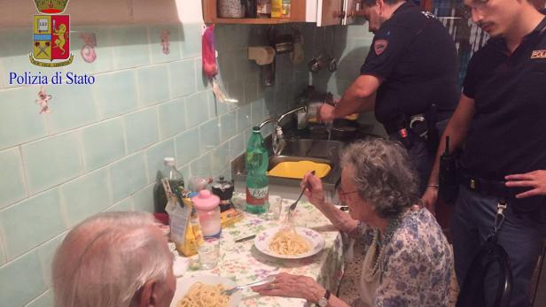 Zwei ältere Menschen essen Spaghetti, während Polizisten in der Küche stehen.