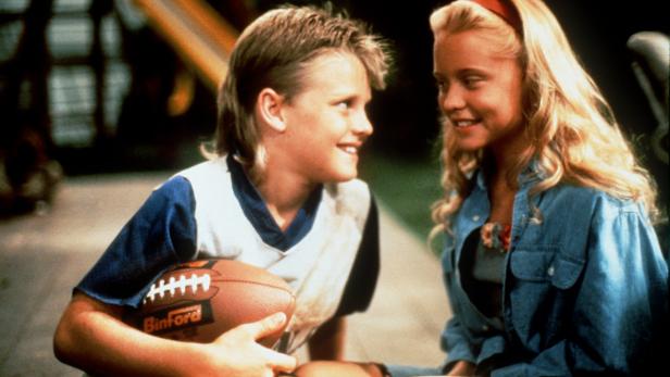 Ein lächelnder Junge mit einem Football in der Hand schaut ein blondes Mädchen an.