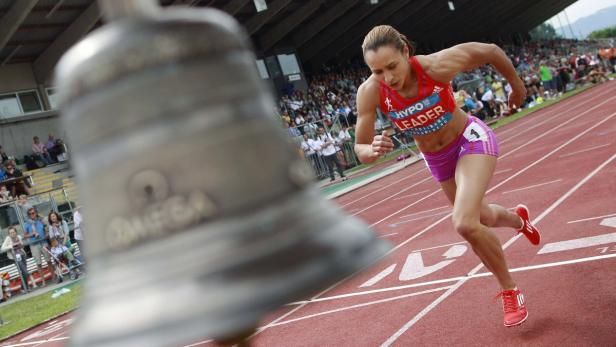 Eine Athletin läuft auf einer roten Tartanbahn, im Vordergrund eine Glocke.