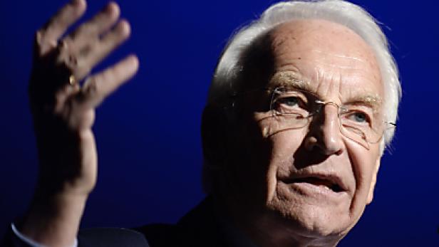 Ein älterer Mann mit Brille gestikuliert vor einem blauen Hintergrund.