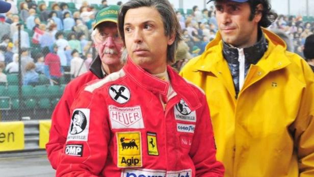 Zwei Männer in Rennfahrerjacken und ein Mann mit gelber Regenjacke stehen vor einer Zuschauertribüne.