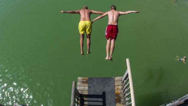 Zwei Männer springen von einem Steg in grünes Wasser.