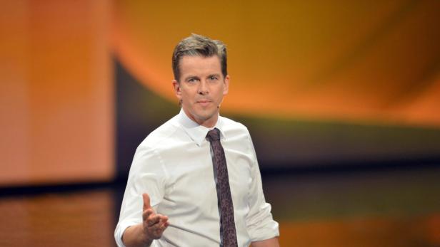 Ein Mann mit weißem Hemd und Krawatte gestikuliert vor einem orangefarbenen Hintergrund.