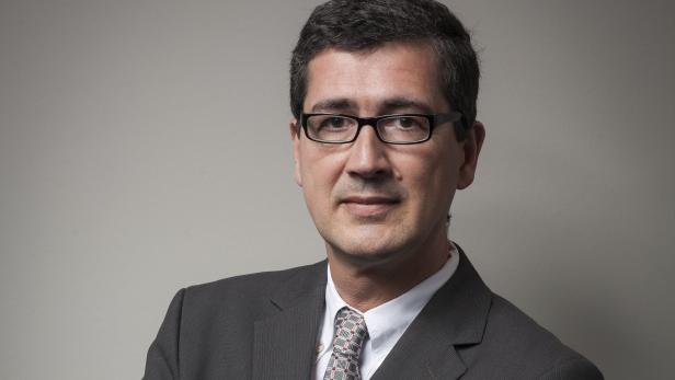 Ein Mann mit Brille und Anzug vor einem grauen Hintergrund.