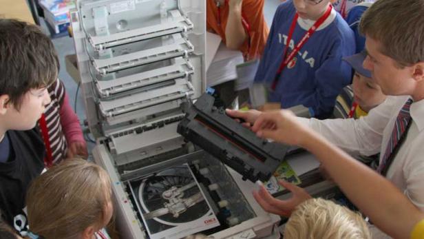 Eine Gruppe von Kindern beobachtet, wie ein Mann eine Tonerkartusche in einen Drucker einsetzt.