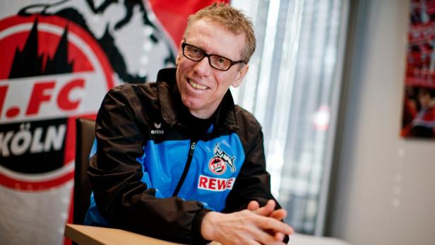 Ein Mann mit Brille und einer Jacke des 1. FC Köln sitzt an einem Tisch.