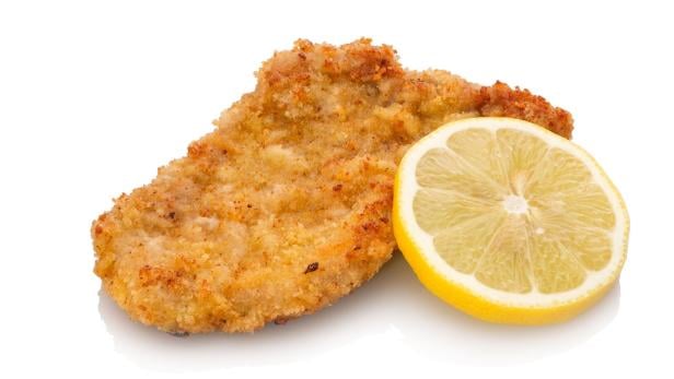 Veal escalope with panade and lemon Ein paniertes Schnitzel liegt neben einer halbierten Zitronenscheibe auf weißem Hintergrund.