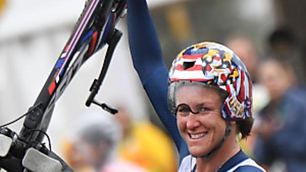 Eine Triathletin im USA-Trikot hält ihr Fahrrad hoch und lächelt.