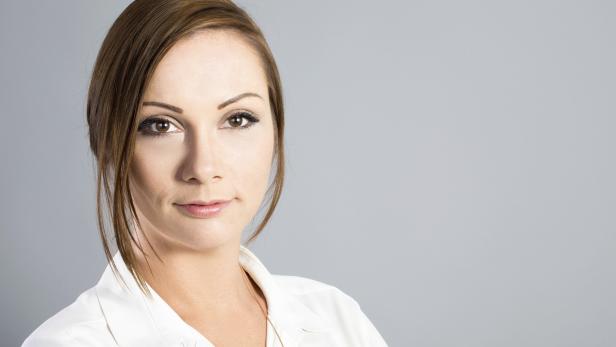 Porträt einer Frau mit braunen Haaren und einem weißen Hemd vor grauem Hintergrund.