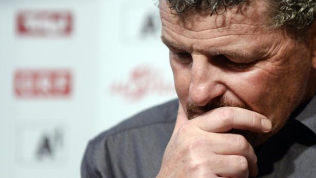 Ein Mann mit Schnurrbart blickt nachdenklich nach unten und berührt sein Kinn.