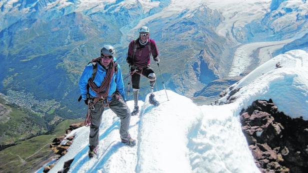 Zwei Bergsteiger stehen auf einem schneebedeckten Gipfel mit Blick auf eine Berglandschaft.