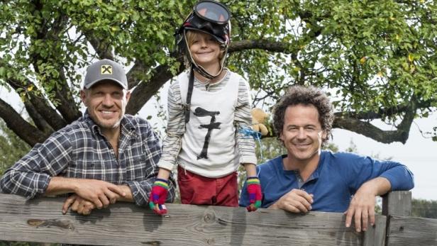 Zwei Männer und ein Kind mit Fliegerhelm lehnen an einem Holzzaun.