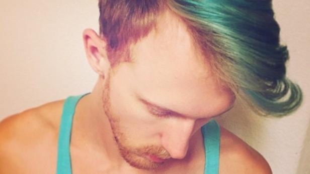 Ein Mann mit einem türkisfarbenen Tanktop und teilweise türkisfarben gefärbten Haaren blickt nach unten. Auf seiner Brust befindet sich ein Tattoo.