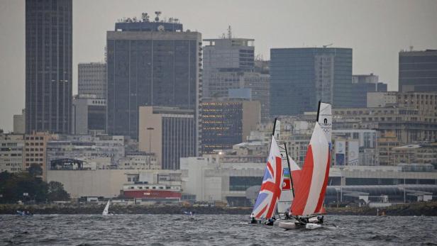 Mehrere Segelboote mit verschiedenen Nationalflaggen vor der Skyline einer Stadt.