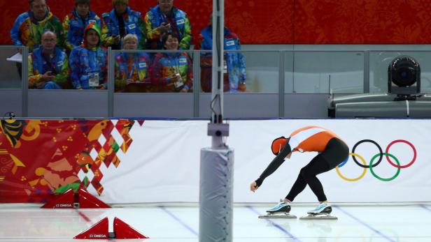 Eine Eisschnellläuferin in oranger Wettkampfkleidung auf einer Eisbahn mit den olympischen Ringen.