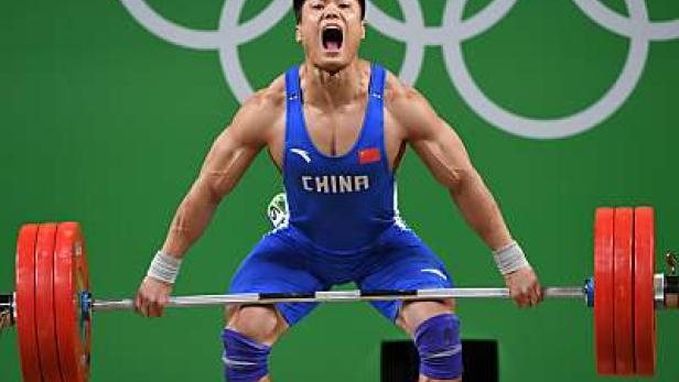 Ein chinesischer Gewichtheber stemmt eine Hantel vor dem Hintergrund der olympischen Ringe.