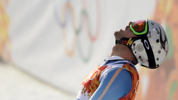Ein Skirennläufer blickt mit Helm und Skibrille nach oben, im Hintergrund die olympischen Ringe.