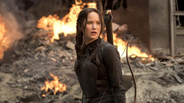 Jennifer Lawrence als Katniss Everdeen vor einem zerstörten Gebäude und Flammen.