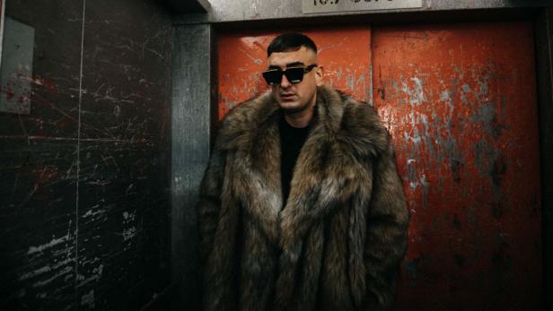 Haftbefehl posiert in einem Pelzmantel und einer Sonnenbrille vor einer roten Tür.