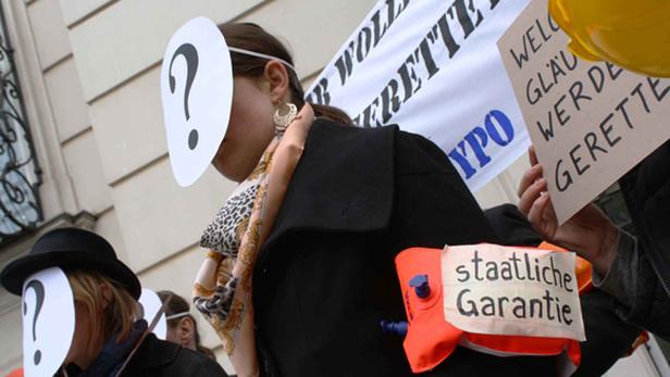 Personen demonstrieren mit Fragezeichenmasken und Schildern, die „staatliche Garantie“ fordern.
