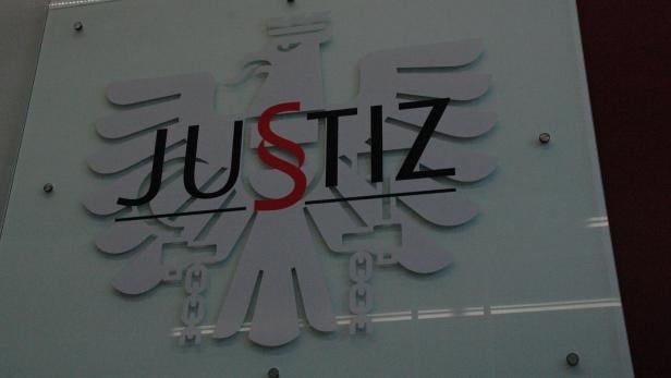 Das Logo der Justiz mit Adler und dem Wort "Justiz", wobei das "S" rot ist, ist an einer Wand befestigt.