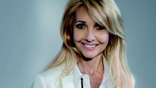 Eine lächelnde Frau mit blonden Haaren und weißem Blazer vor einem hellblauen Hintergrund.
