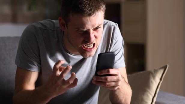 Ein wütender Mann sitzt auf einem Sofa und starrt auf sein Smartphone.