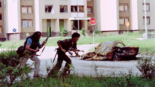 Zwei bewaffnete Männer ducken sich hinter einem ausgebrannten Auto vor einem von Einschusslöchern übersäten Wohnhaus.