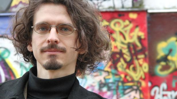 Ein Mann mit Brille, Bart und lockigem Haar vor einer Wand mit Graffiti.