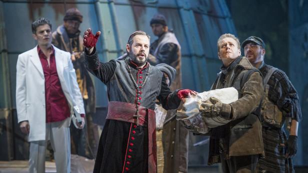 Eine Theaterszene mit einem Priester in roter Kleidung und mehreren anderen Darstellern.