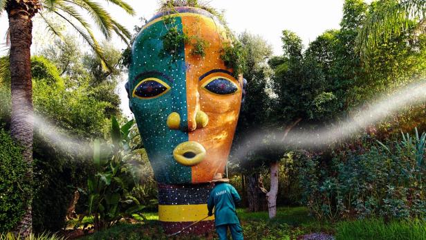 Ein Gärtner bewässert eine große, mosaikartige Gesichtsstatue in einem üppigen Garten.