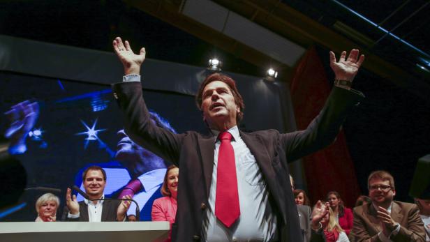 Ein Mann in Anzug und roter Krawatte steht mit erhobenen Armen vor Publikum.