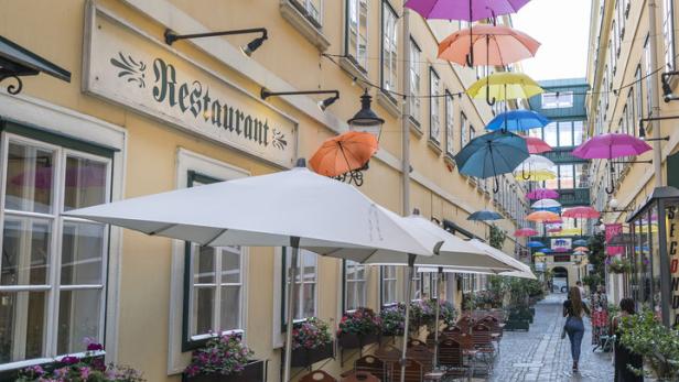 Eine Gasse mit einem Restaurant und bunten, schwebenden Regenschirmen.